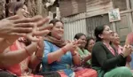 A group of South Asian women in colourful clothes sit on the floor smiling and clapping